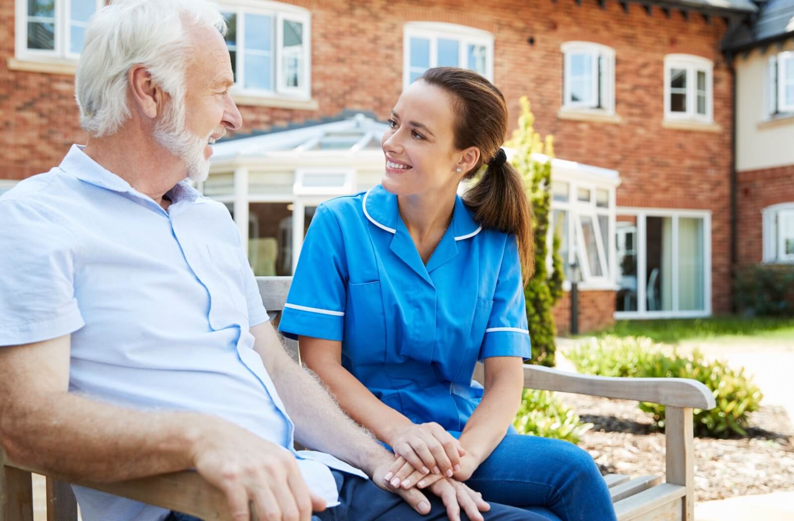An older adult man talks happily on a bench outside with a staff member in an assisted living community.
