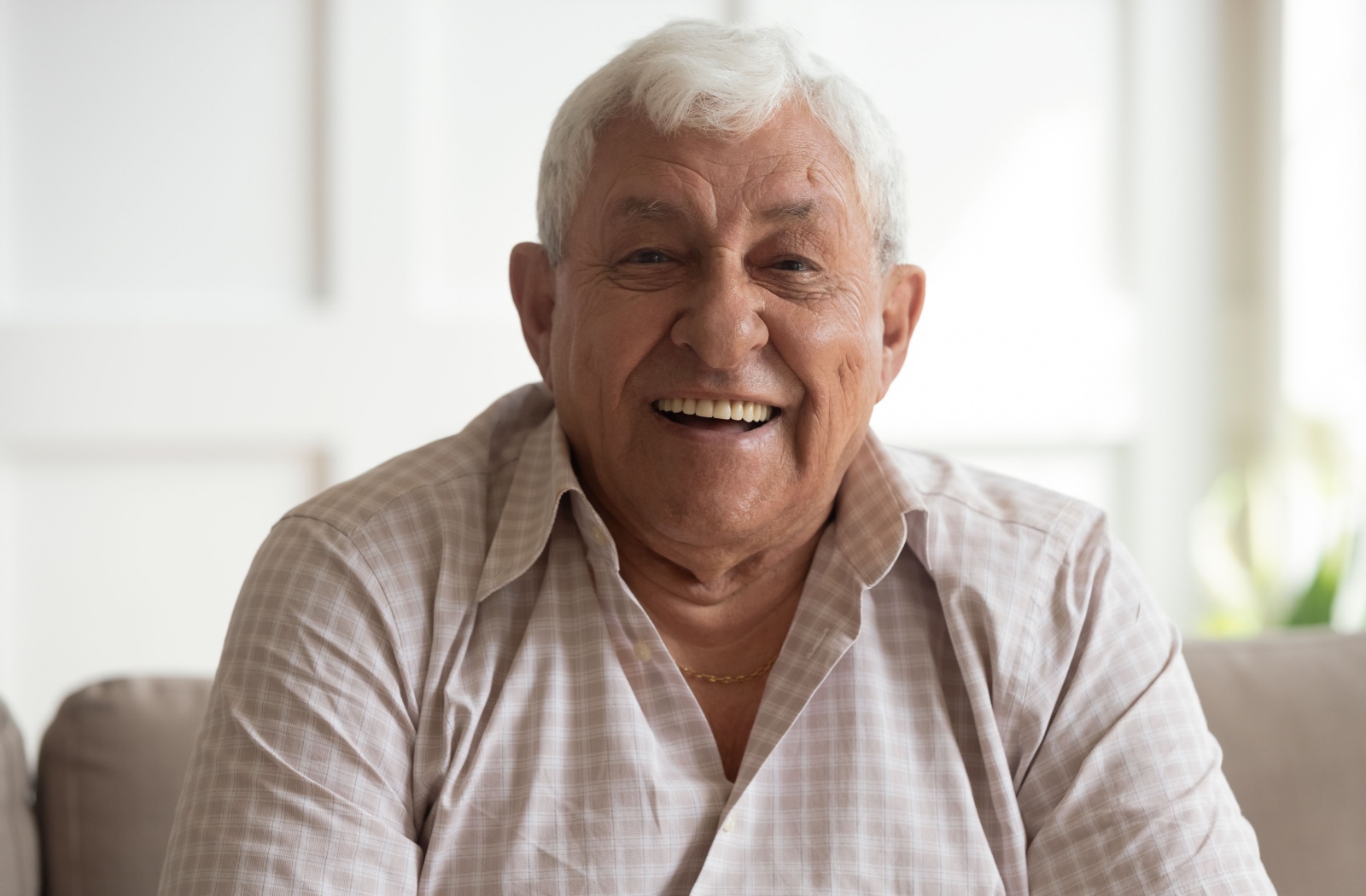 An older adult man laughs as he sits on a couch.