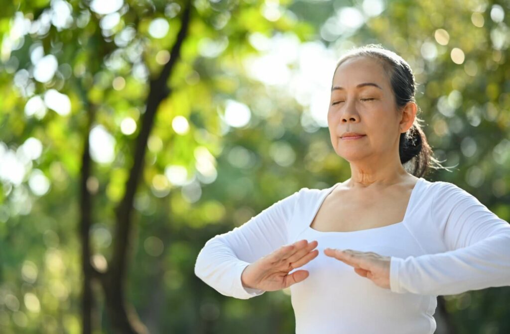 A calm older adult woman closes her eyes as she practices mindfulness during tai chi in nature.