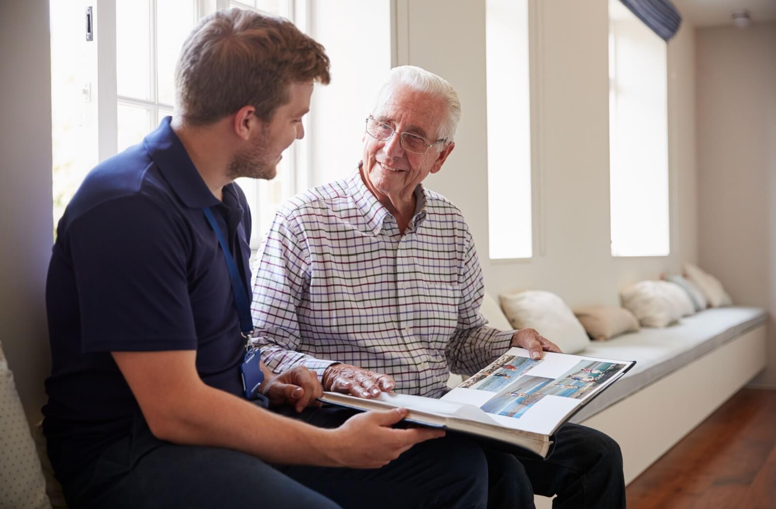 An older adult man and a memory care staff flipping through a photo album in a sunlit space.