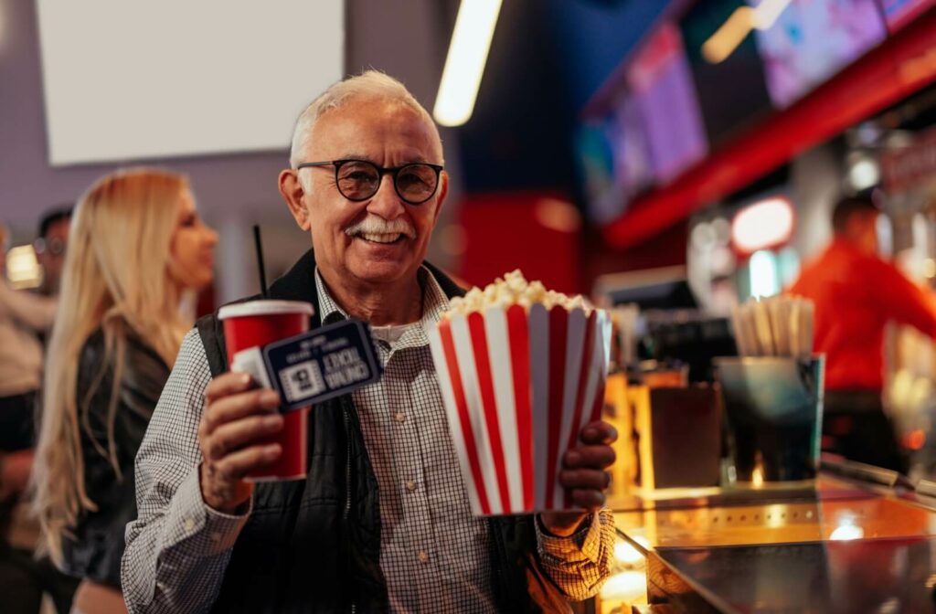 A smiling older adult holds popcorn and a drink at a movie theatre's concession stand.