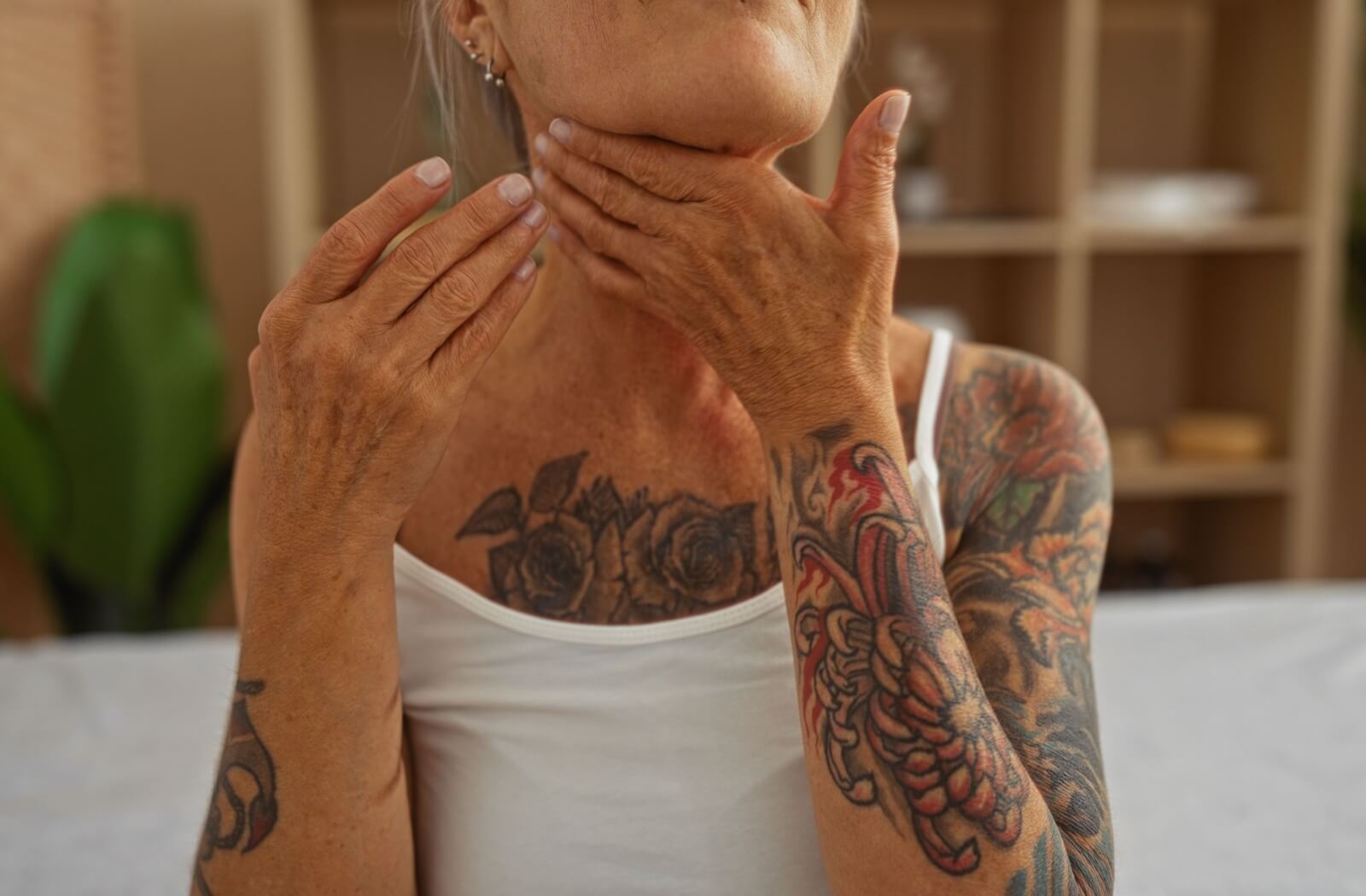 A tattooed older adult moisturizes their skin, rubbing their neck with their hand.