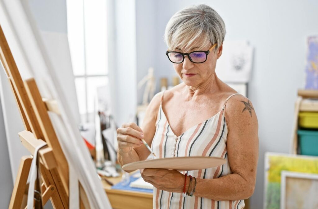 An older adult with a shoulder tattoo works on a painting in an art studio.