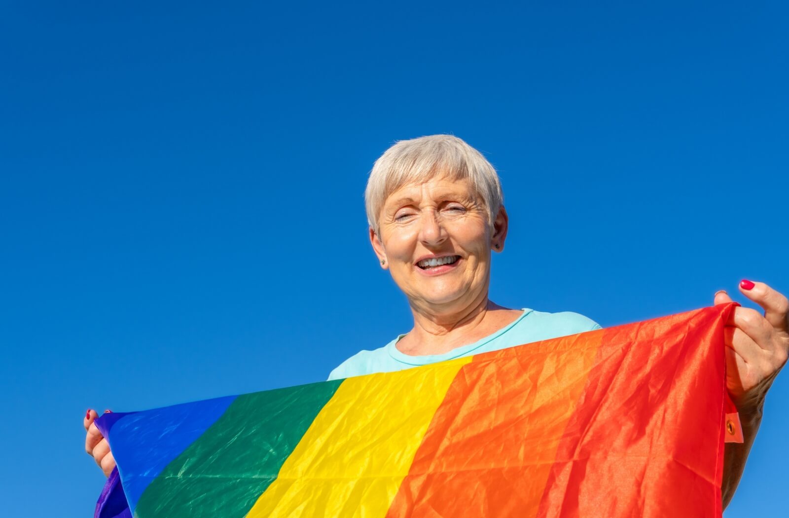 An LGBTQ older adult holds up a pride flag while smiling against a clear, deep blue sky.