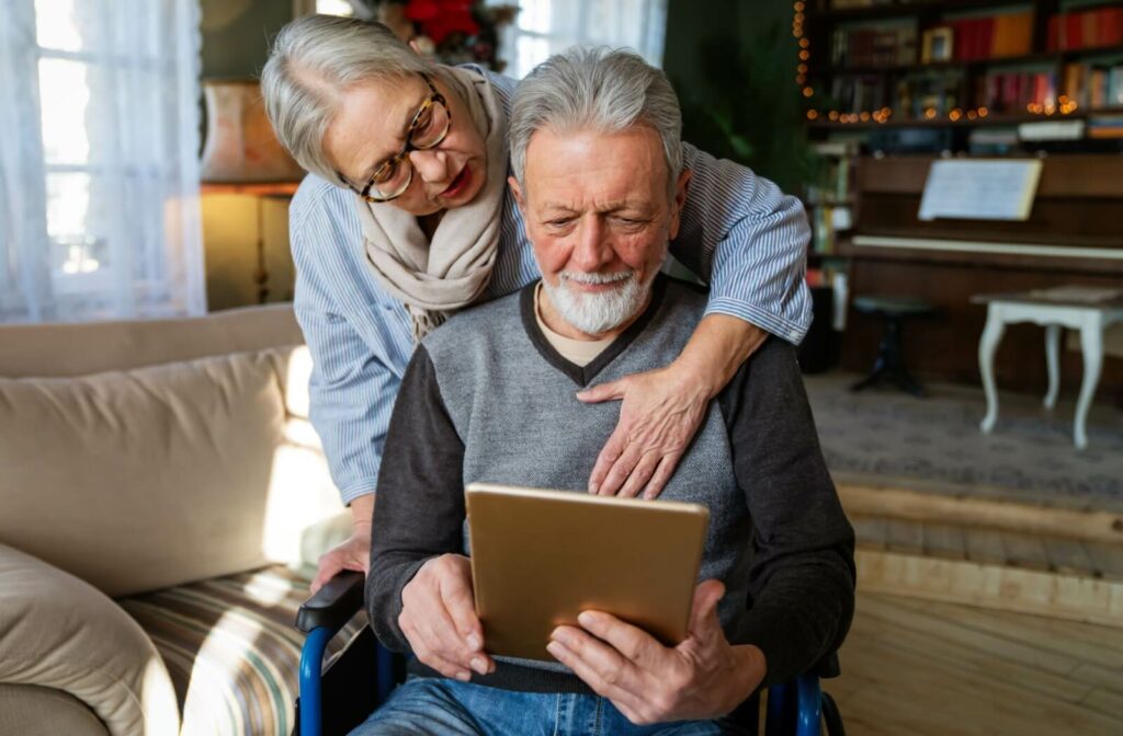 An older adult hugging their spouse in a wheelchair from behind while they use a tablet to take a virtual tour together.
