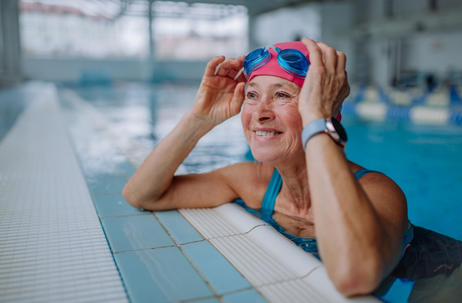 A smiling older adult in swim gear adjusting goggles at the edge of an indoor pool.