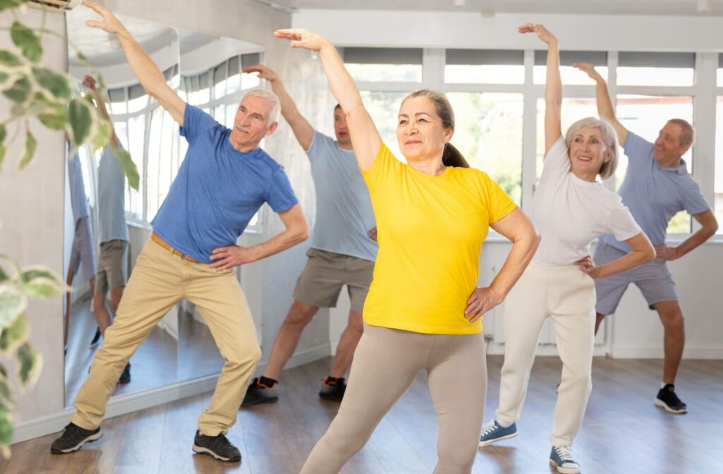 Group of older adults doing standing side stretches together in a bright fitness studio.