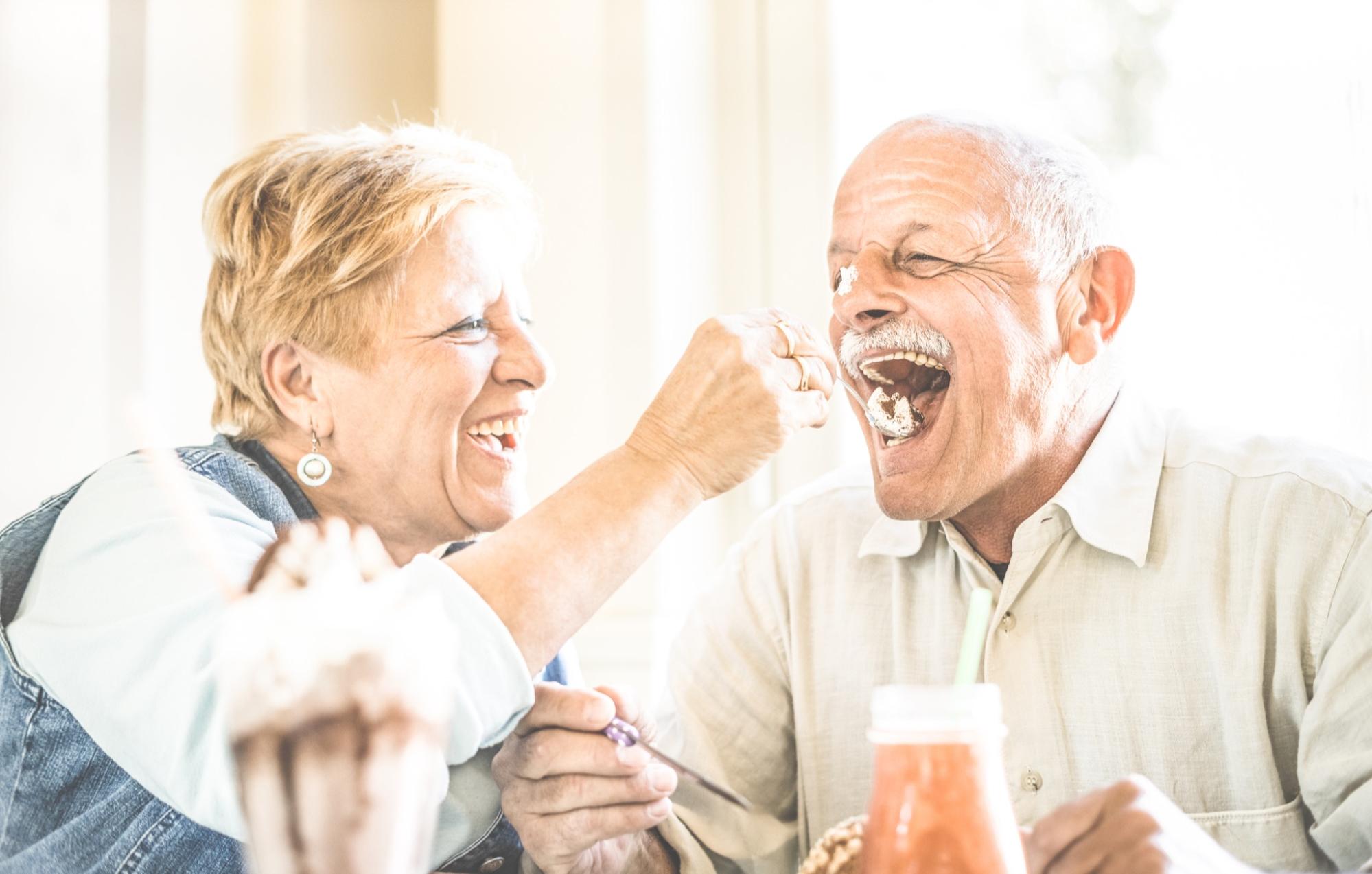 A happy, retired older couple eating together. The woman is putting a spoon of food in her husband's open, smiling mouth.
